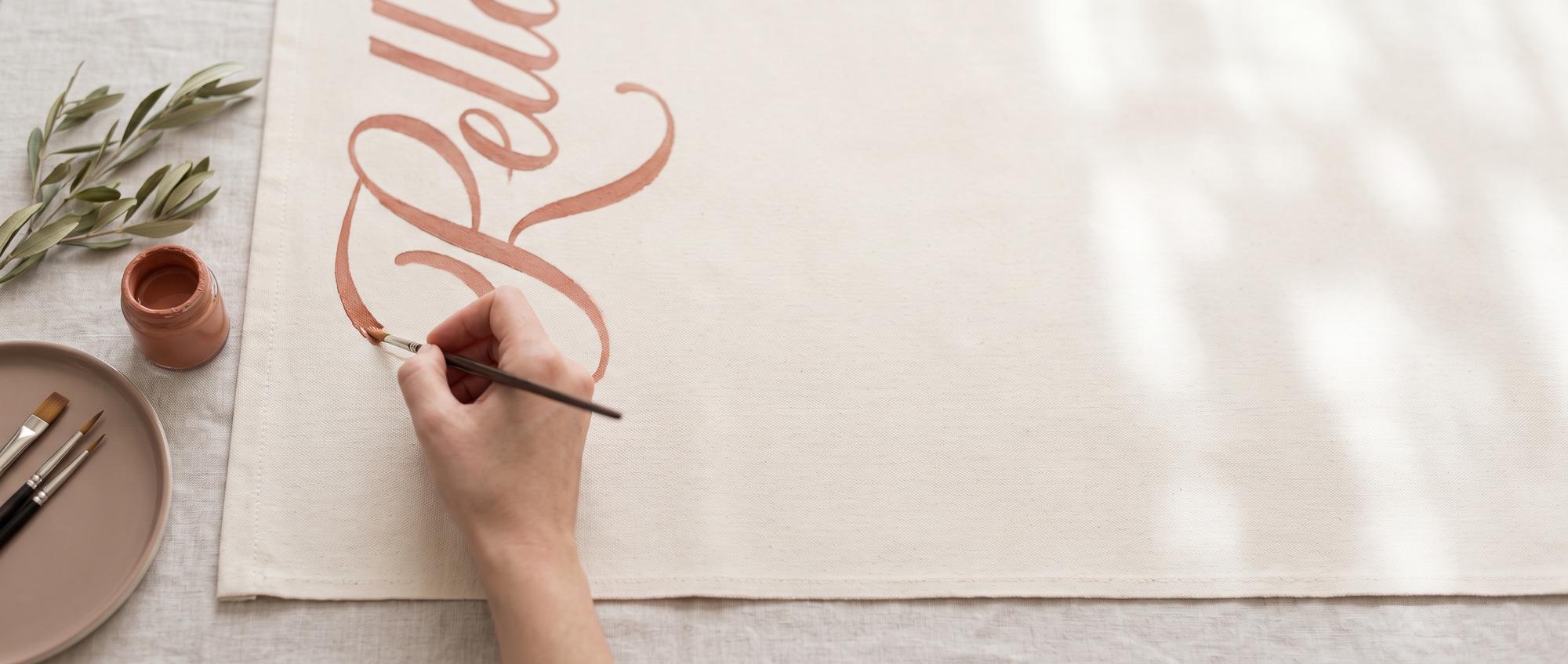 A hand painting a flowing terracotta letterform onto cream canvas, beside a jar of paint and olive sprigs
